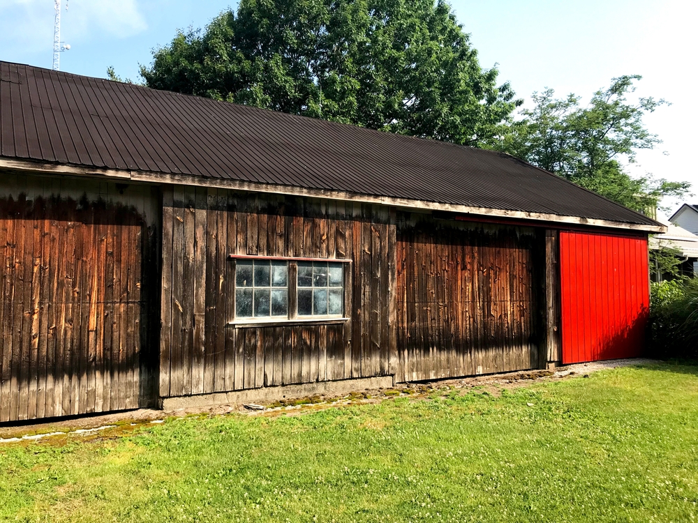 Old weathered country outbuilding. Garage work shop man cave. Wood wooden board boards siding. Red. Multi-paned window. Barn doors. Green grass, Trees.